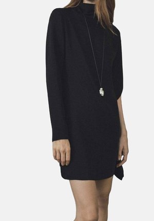 Femme portant une mini-robe noire à manches longues avec un col montant et un long collier avec pendentif.