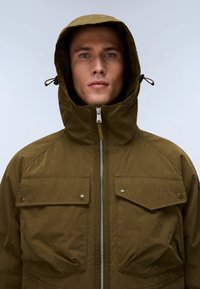 Άνδρας που φοράει χακί πράσινο μπουφάν με κουκούλα, φερμουάρ μπροστά και κουμπωτές τσέπες στο στήθος, στραμμένος προς τα εμπρός μπροστά σε μονόχρωμο φόντο.