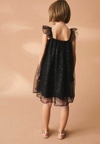 Robe en tulle noire avec des manches froncées, ornée de sequins argentés éparpillés. Forme A-line, matériau léger et design à dos ouvert.