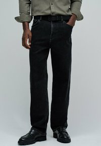 Hombre vestido con jeans negros, cinturón negro, mocasines negros y una camisa verde oliva abotonada con las mangas arremangadas.