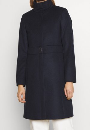 Nainen pukeutuneena tummansiniseen villakangastakkiin, jossa on korkea pystykaulus, piiloketjullinen etuosuus ja vyötäröllä saumadetalji, yhdistettynä valkoisiin housuihin.