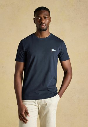 Mand iført en mørkeblå t-shirt med et lille lysblåt og hvidt flagemblem på brystet og beige bukser, der står mod en ensfarvet baggrund.