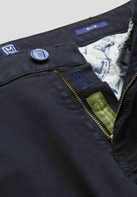 Pantalon slim-fit bleu marine avec une texture lisse, présentant un bouton bleu, une fermeture éclair dorée et un motif interne. Les étiquettes indiquent qu'il est fabriqué en Europe.