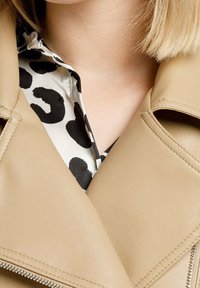 Giacca in pelle beige con colletto ampio, caratteristiche di cuciture visibili e chiusura con zip. Sotto, una camicia bianca con motivi astratti neri.