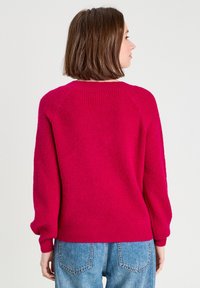 Suéter ajustado de color rosa brillante con textura acanalada, mangas raglán y puños ajustados, combinado con jeans de color azul claro. Vista trasera mostrada.