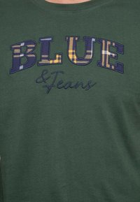 T-shirt vert olive avec un texte brodé multicolore "BLUE & Jeans", agrémenté d'un motif à carreaux dans des nuances de bleu, jaune et violet.