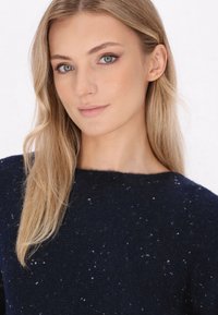 Dunkelblauer Strickpullover mit Rundhalsausschnitt und strukturierter Oberfläche, die durch kleine weiße Sprenkel im Stoff ergänzt wird.
