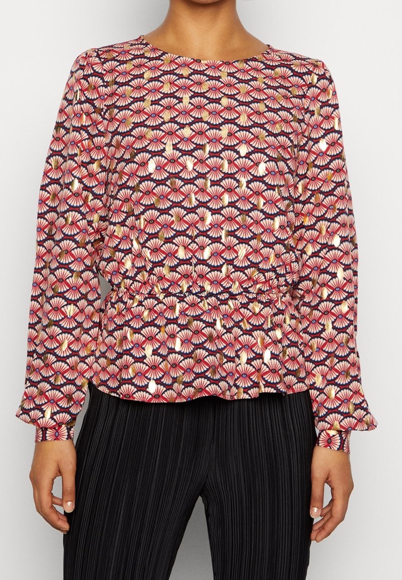 Femme portant un chemisier à manches longues avec des motifs éventail roses, bleus marine et dorés, associé à un pantalon plissé noir.