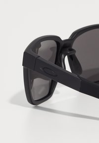 Ochelari de soare negri cu un finisaj mat, lentile rectangulare și rame groase. Caracteristicile includ un logo pe latură și brate laterale curbate.