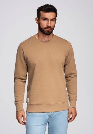 Felpa beige a girocollo realizzata in tessuto morbido, con maniche lunghe e orlo a costine. Indossata con jeans azzurri chiari.