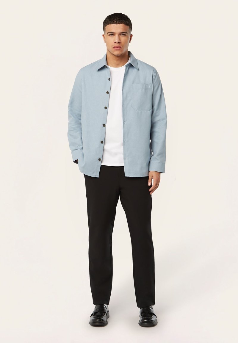 Lichtblauw, langmouwen shirt met knoopsluiting, een borstzak en donkere knopen, gecombineerd met zwarte broek en schoenen.