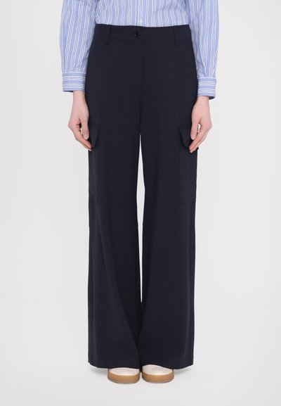 Pantaloni cargo larghi di colore blu navy con tasche laterali, una texture liscia e chiusura con bottone in vita, abbinati a scarpe beige slip-on.
