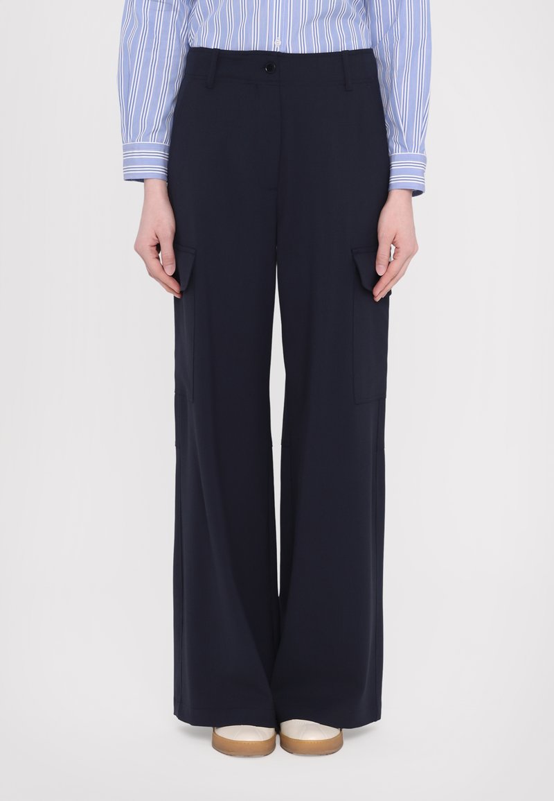 Pantalons cargo larges navy avec des poches latérales, une texture lisse et une fermeture à bouton à la taille, associés à des chaussures beiges à enfiler.