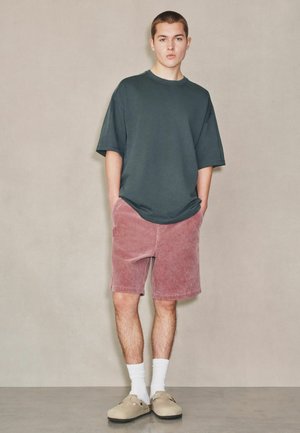 Young man standing with hands in pockets wearing dark oversized t-shirt, pink shorts, white socks, and beige slip-on shoes against plain background.