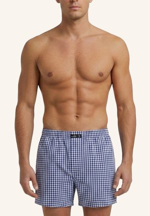 Hemdloser männlicher Model trägt blau-weiß karierte Boxershorts und steht mit entspannten Armen vor schlichtem Hintergrund.
