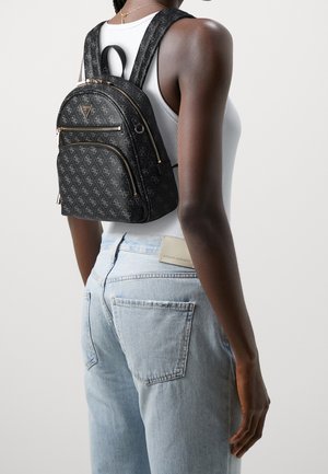 Femme portant un haut blanc sans manches et un jean bleu clair avec un petit sac à dos de créateur noir orné de fermetures éclair et de logos dorés.