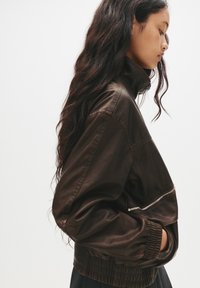 Giacca bomber in pelle marrone con colletto alto, polsini elasticizzati e dettaglio con tasca con zip. Finitura testurizzata, vestibilità rilassata e design cropped.