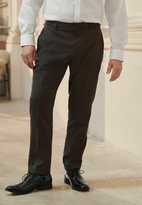 Pantalons marron en tissu lisse, dotés d'une coupe ajustée, de poches latérales et portés avec des chaussures noires à lacets brillantes.
