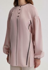 Lichtroze blouse met een ronde halslijn, zwarte knoopsluiting en decoratieve stiksel langs de zijkanten. Heeft pofmouwen en splitten.
