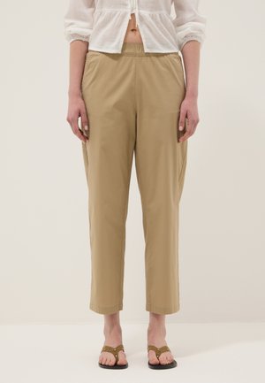 Donna con pantaloni beige a vita elastica e gamba dritta, camicetta corta bianca e sandali marroni con dettagli ritagliati circolari.