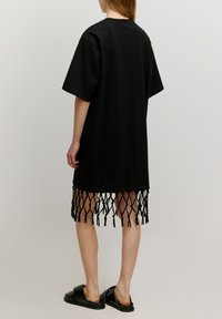 Schwarzes, überdimensionales T-Shirt-Kleid mit einem geknoteten Fransen-Saum, aus weichem Stoff gefertigt, mit kurzen Ärmeln und einer lockeren Passform. Getragen mit schwarzen Sandalen.