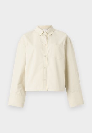 Kuttet, lys beige skjorte laget av bomull, med spiss krage, frontknapping og en enkelt brystlomme med en subtil logo.