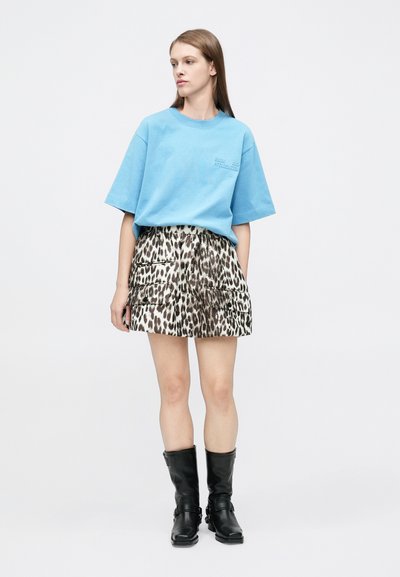 Femme portant un t-shirt bleu ample, une mini-jupe imprimé léopard avec poches et des bottes noires mi-mollet, debout devant un fond blanc uni.