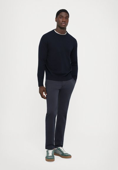 Homme portant un pull bleu marine, un pantalon gris foncé et des baskets vertes, se tenant avec une main dans la poche contre un fond blanc.