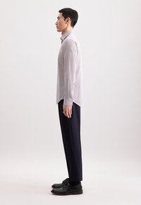 Camicia a righe grigio chiaro con maniche lunghe, dettagli blu al polsino. Abbinata a pantaloni scuri e scarpe nere. Vista laterale, postura eretta.