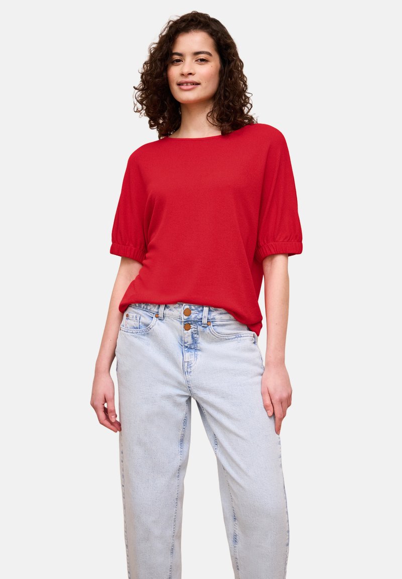 Femme aux cheveux bouclés et foncés, portant un chemisier rouge à manches courtes rentré dans un jean bleu clair taille haute, devant un fond uni.