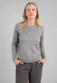 Donna con capelli biondi che indossa un maglione grigio a maniche lunghe e pantaloni abbinati, in piedi con le mani in tasca su uno sfondo neutro.