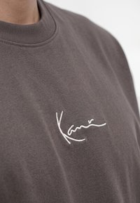 Nahaufnahme einer Person, die ein braunes Hemd mit rundem Kragen und weißem gesticktem Text auf der Brust trägt.