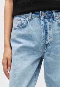 Ανοιχτό μπλε τζιν με χαλαρή εφαρμογή, με χαλκομανίες, πέντε τσέπες και διακριτική ξεθώριαση. Το ύφασμα έχει απαλή υφή.