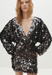 Musta ja hopeanvärinen paljettinen mekko, jossa on syvä v-pääntie ja pitkät, pöhöttyneet hihat. Lyhyt pituus, peitetty pyöreillä paljeteilla, jotka luovat heijastavan tekstuurin.