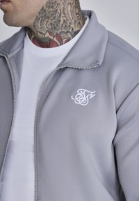 Giacca grigia chiaro con zip e colletto alto, logo ricamato bianco sul lato sinistro del petto e una maglietta bianca sotto. Tessuto dalla superficie liscia.
