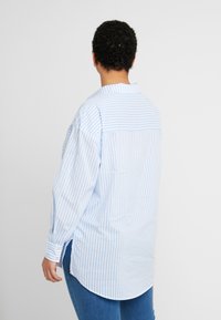 Camisa de manga larga a rayas azul y blanca con puño abotonado y cortes laterales, que presenta un yugo trasero y un dobladillo redondeado.