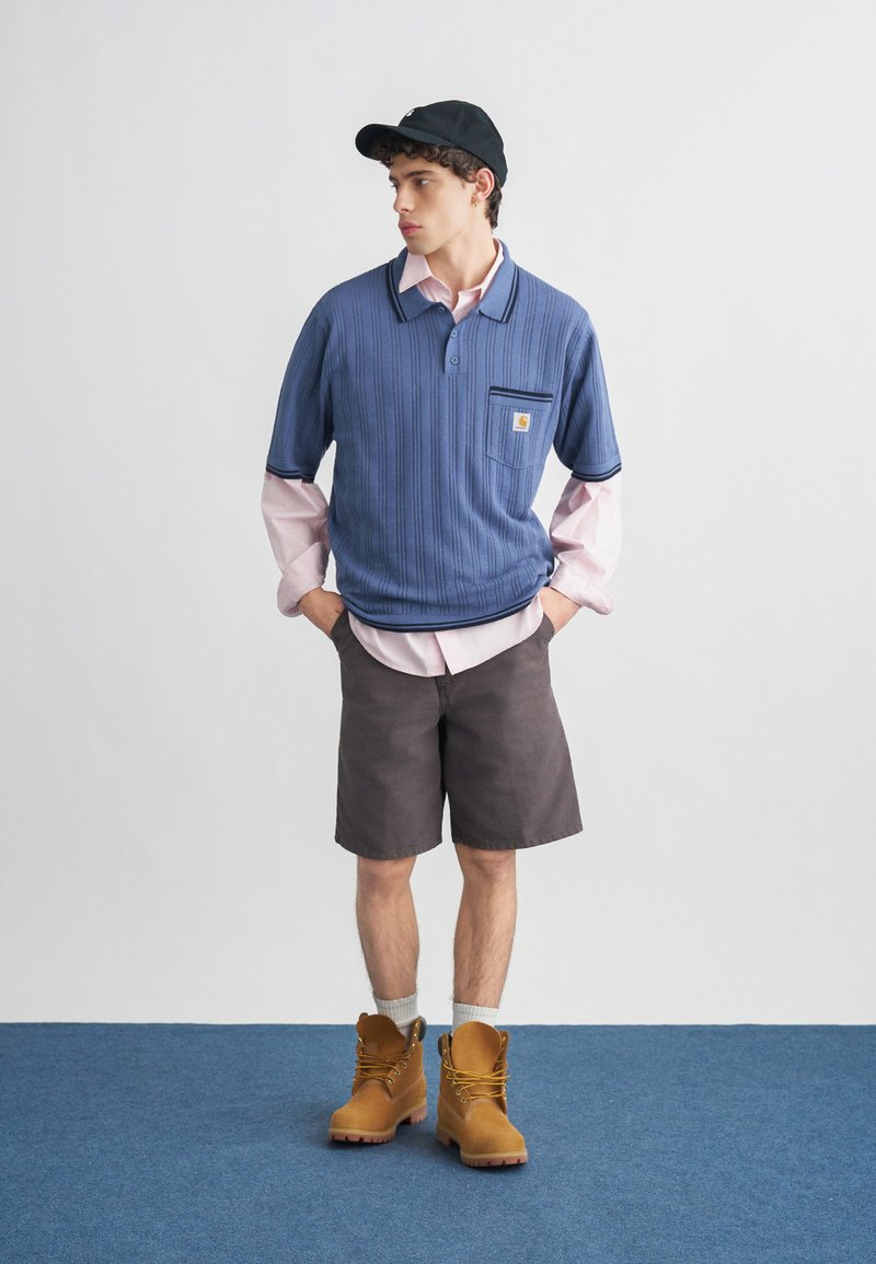 Jonge man die een blauw kortärmelig truitje draagt over een roze overhemd, donkere korte broek, beige laarzen en een zwarte pet, staand op een blauw tapijt tegen een witte muur.