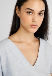 Goldene Anhängerkette mit einem "S" Charm, getragen an einer feinen Kette. Das Model trägt ein hellblaues V-Ausschnitt-Oberteil aus einem strukturierten Stoff.