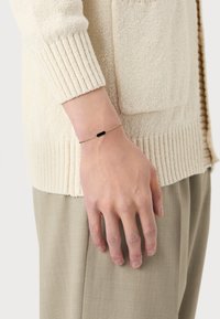 Goldene Kettenarmband mit einem schwarzen rechteckigen Stein, getragen an einer Hand gegen einen cremefarbenen strukturierten Pullover, in einem entspannten, neutralen Outfit.