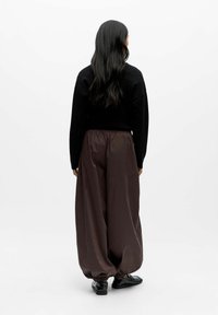 Un pull noir associé à un pantalon lâche de couleur marron foncé et brillant, doté d'une taille élastique et de poignets froncés, porté avec des chaussures noires.
