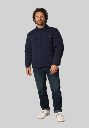Veste matelassée bleu marine avec un col de chemise, deux poches sur la poitrine et des poignets élastiques, assortie à un jean foncé et des baskets blanches.