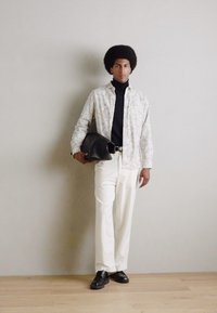 Camisa de manga larga con estampado, suéter negro de cuello alto, pantalones blancos y zapatos negros. Sosteniendo un bolso negro, de pie contra un fondo neutro.