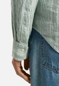 Camicia a maniche lunghe verde e bianca a righe con bottoni; dettaglio della mano visibile e jeans in denim con tasca, che mostrano la texture del tessuto.