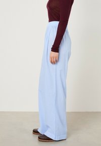 Šviesiai mėlynos dryžuotos plačios kelnės su elastine juosmens dalimi, derinamos su rudos spalvos slip-on batais. Lygus audinys ir laisvas siluetas.