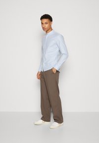Camicia azzurra chiaro con colletto arrotondato, abbinata a pantaloni marroni e sneaker bianche. Tessuto morbido, vestibilità su misura, design minimale.