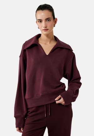 Vrouw draagt bijpassende donker bordeauxrode trui met brede kraag en bijpassende broek met trekkoord, staand tegen een effen lichte achtergrond.