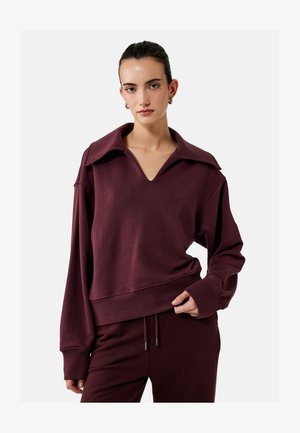 Vrouw draagt bijpassende donker bordeauxrode trui met brede kraag en bijpassende broek met trekkoord, staand tegen een effen lichte achtergrond.