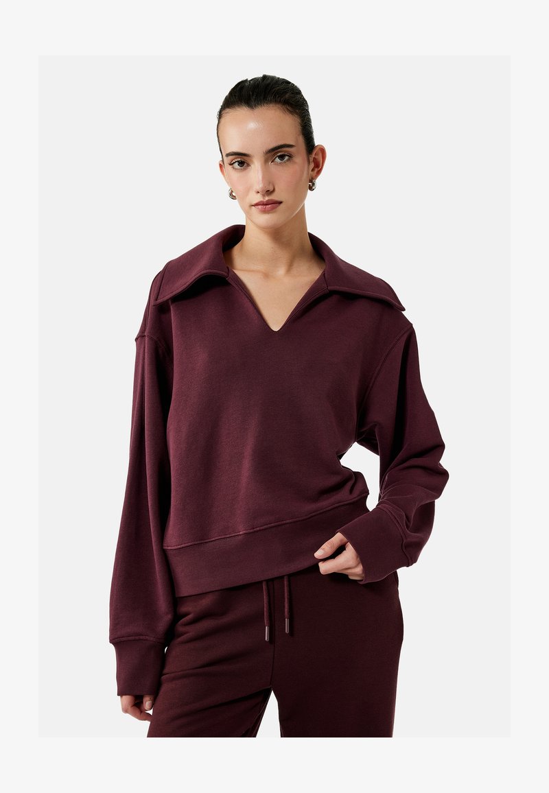 Kvinde iført matchende mørkerød sweatshirt med bred krave og matchende snorbukser, stående mod en ensfarvet lys baggrund.