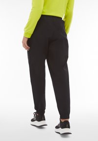 Schwarze Jogginghosen mit elastischem Bund und Bündchen an den Knöcheln. Kombiniert mit einem leuchtend grünen langärmeligen Oberteil und schwarzen Turnschuhen.