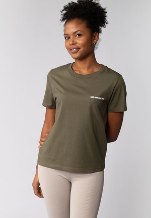 Olive green t-shirt with short sleeves, rounded neckline, and white text detailing; paired with light ribbed leggings. Smooth cotton material.